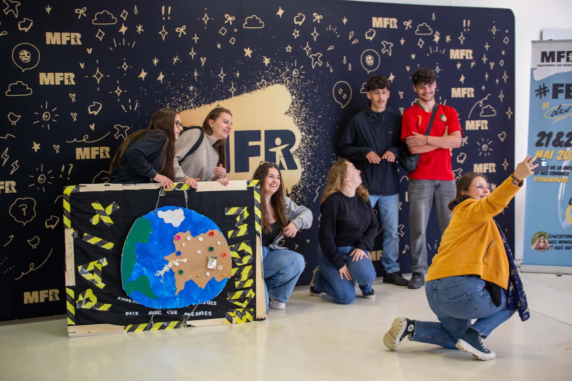 Jeunes posant devant le photocall du Festival des Jeunes MFR à Strasbourg, avec une carte du monde décorative et une animatrice prenant une photo de groupe